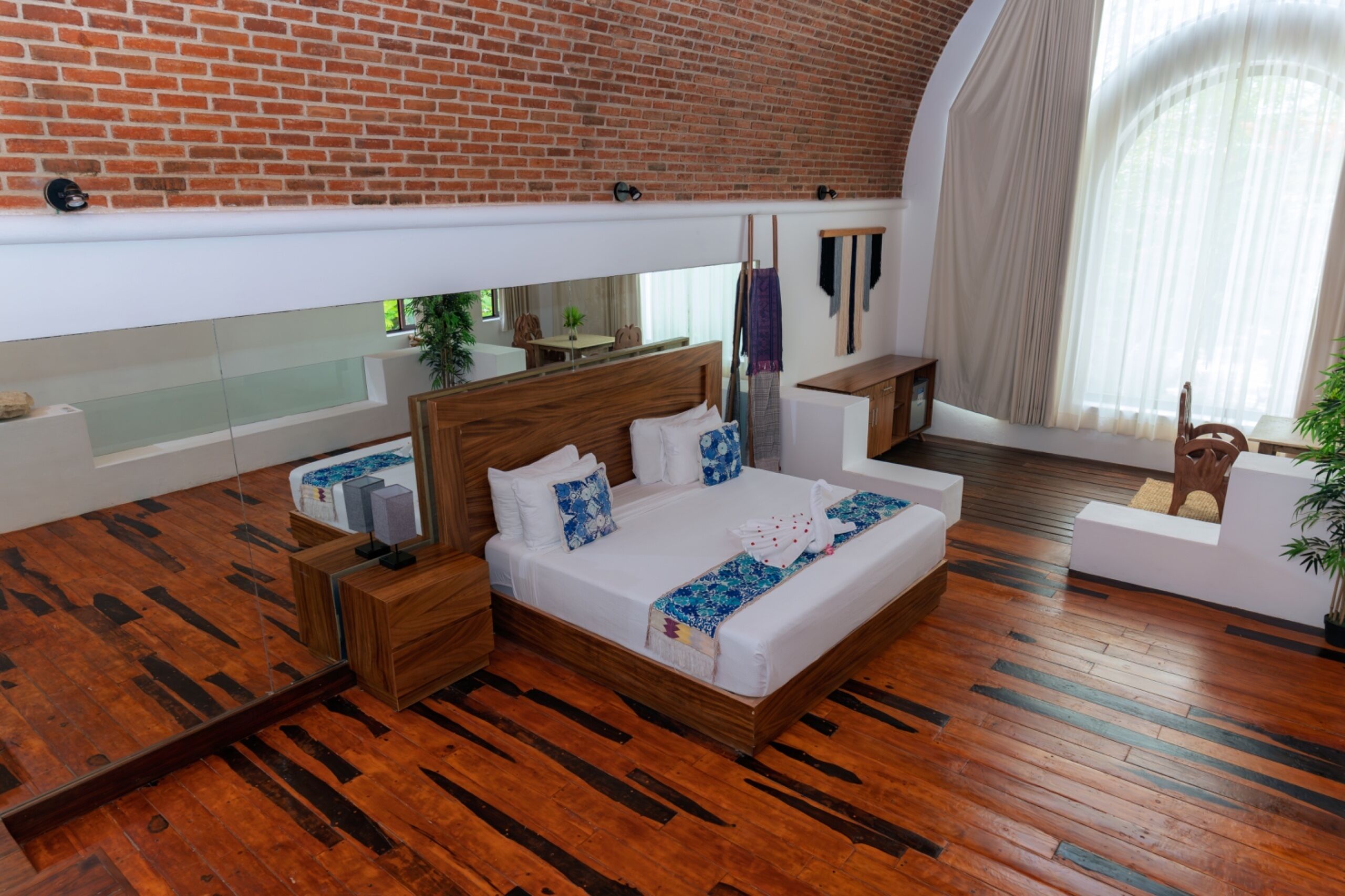 Une chambre d’hôtel moderne avec un mur en briques, un grand lit avec une literie blanche, une tête de lit en bois, un petit coin salon et une grande fenêtre avec des rideaux légers.
