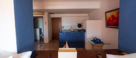 Une pièce avec un plancher en bois, un lit blanc, une petite table avec une lampe, et une vue sur un coin cuisine avec des armoires bleues et un dosseret carrelé.