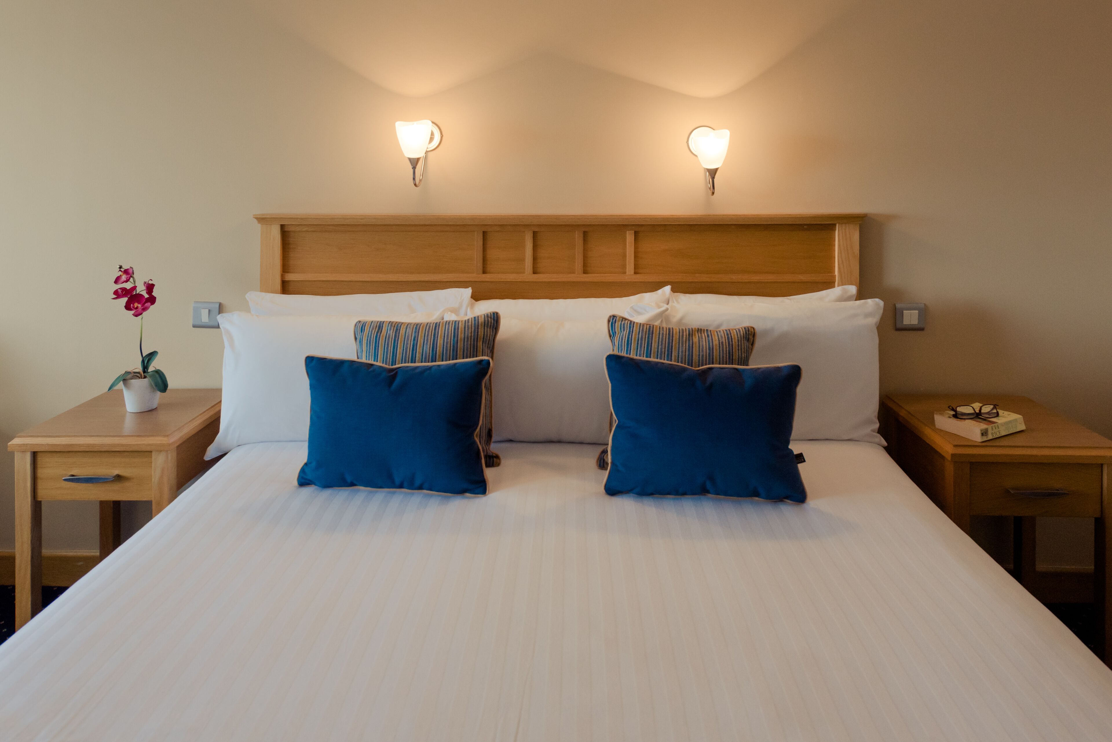 A neatly made bed with two blue pillows and a wooden headboard. There are two bedside tables, one with a plant and the other with a book and glasses.
