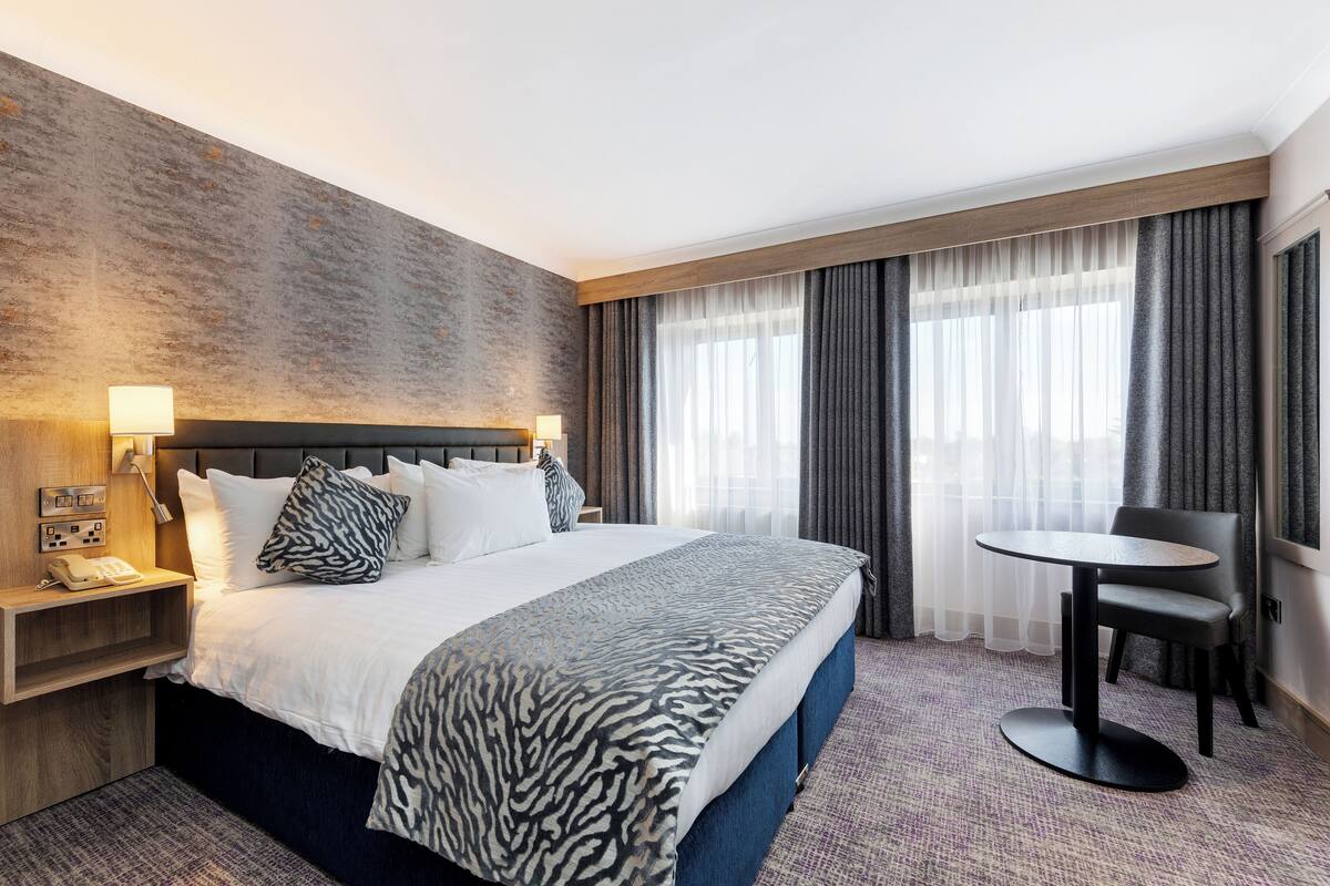 Habitación de hotel con una cama grande, una mesita de noche con lámpara, una pequeña mesa redonda y una silla.