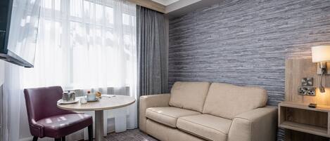Habitación de hotel con un sofá color beige, una mesa redonda con una silla, un televisor montado en la pared y una mesita de noche de madera con una lámpara.