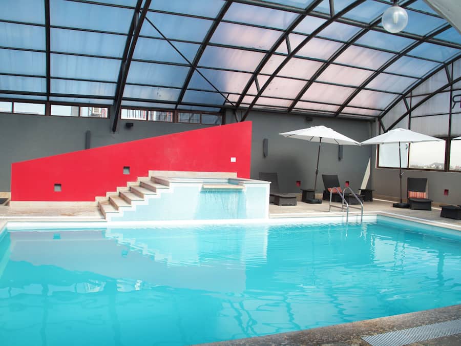 Una piscina interior con una pared roja, escalones que bajan a la piscina y sillones de baño con sombrillas.