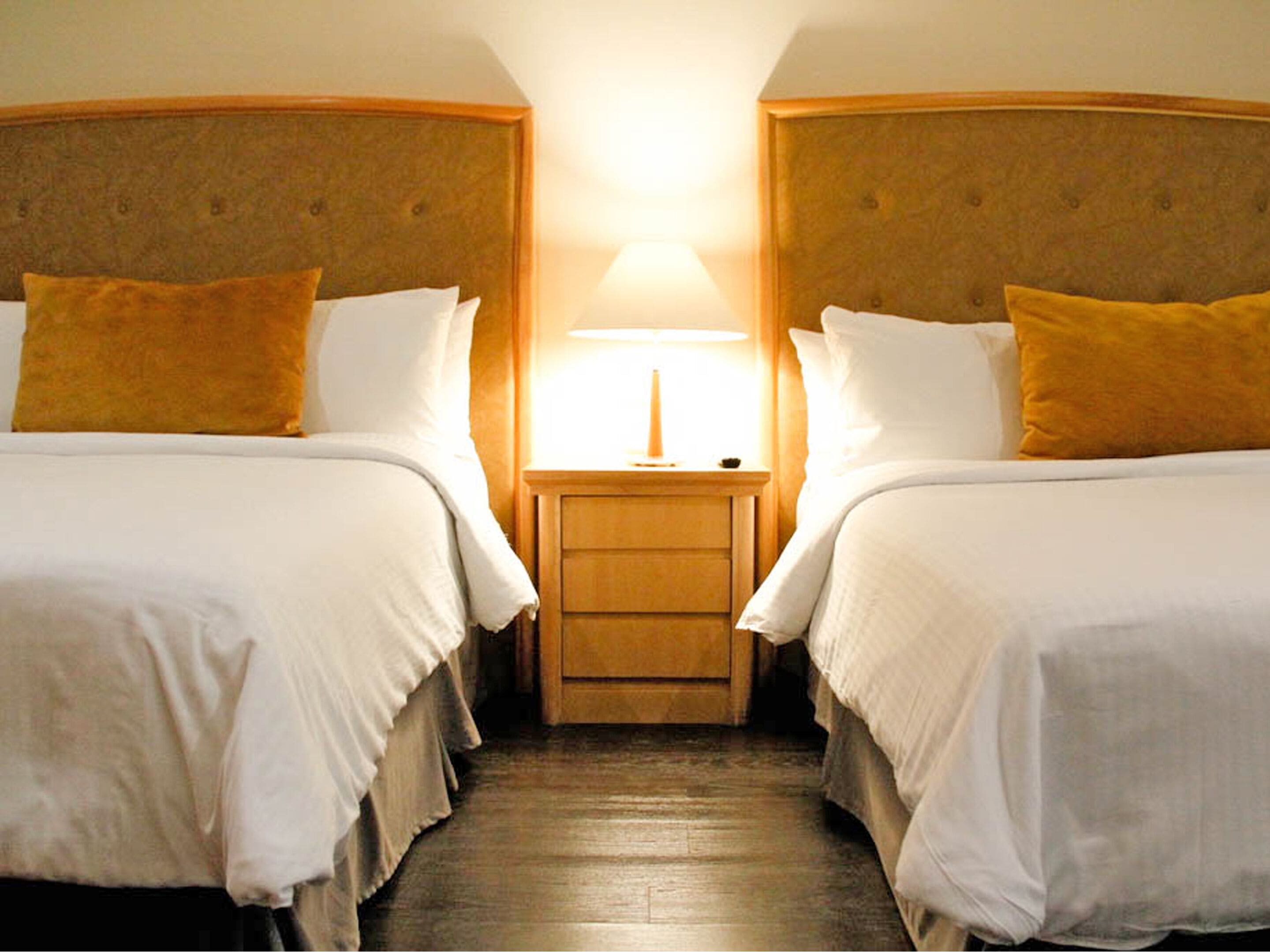 Habitación de hotel con dos camas, una mesita de noche y una lámpara entre ellas.