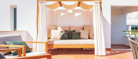 Un espacio al aire libre con luz solar, con un sofá acolchado, sillas de madera y una pequeña mesa redonda.