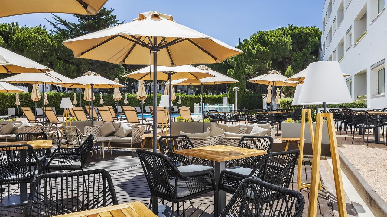 An outdoor seating area with tables and chairs, umbrellas, and a pool in the background.