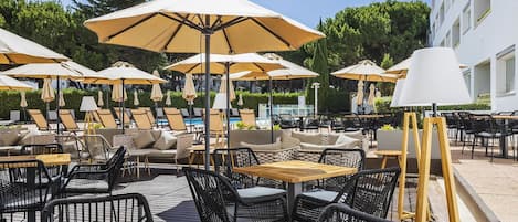 An outdoor seating area with tables and chairs, umbrellas, and a pool in the background.