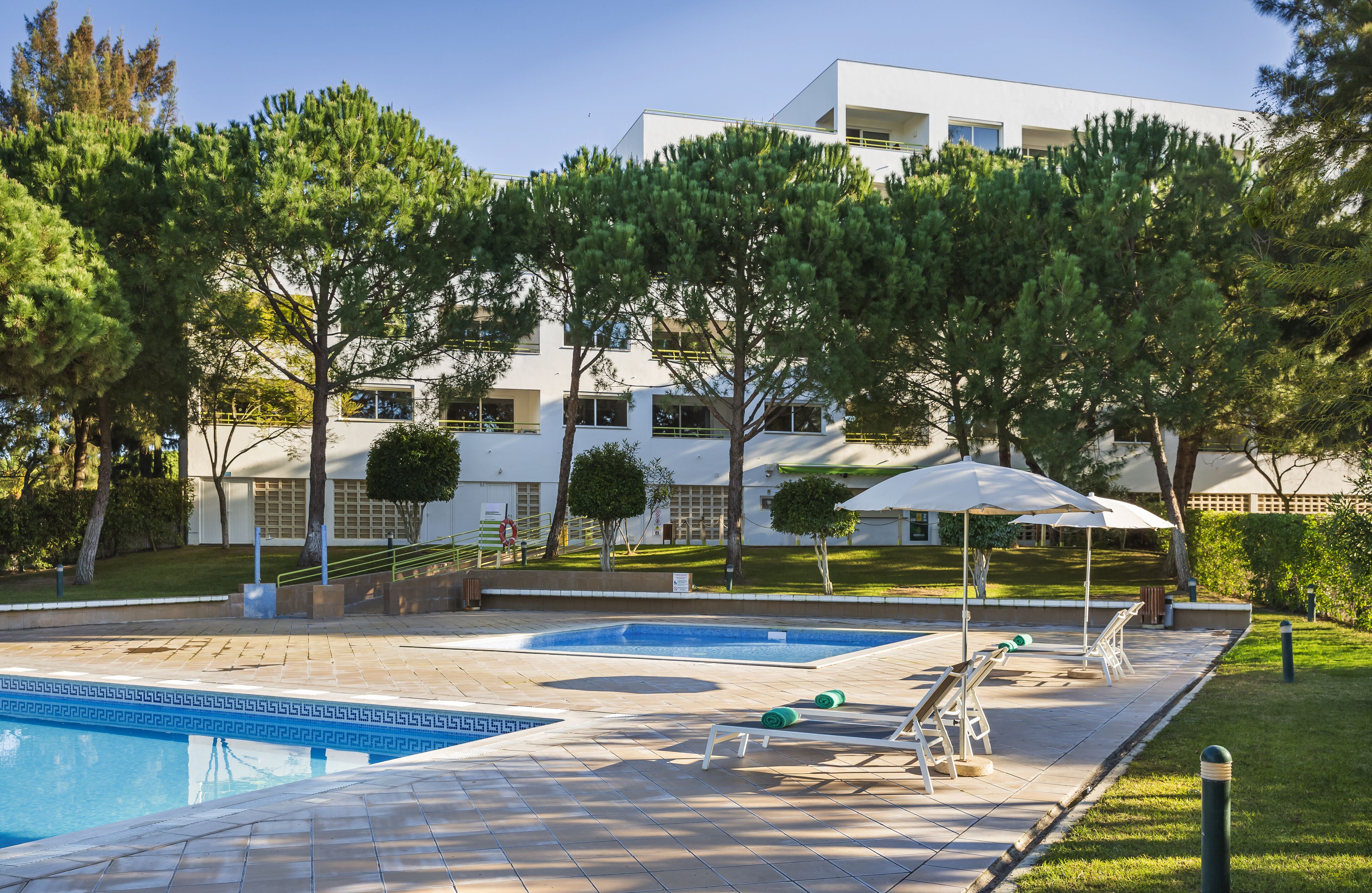 Un espace piscine doté de chaises longues et de parasols, entouré d’arbres et avec un bâtiment moderne en arrière-plan.