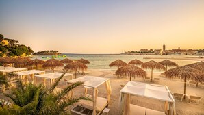 A beachside hotel with thatched umbrellas, white lounge chairs, and a view of the sea and cityscape. - Imperial Heritage Hotel, Valamar Collection (Rab)