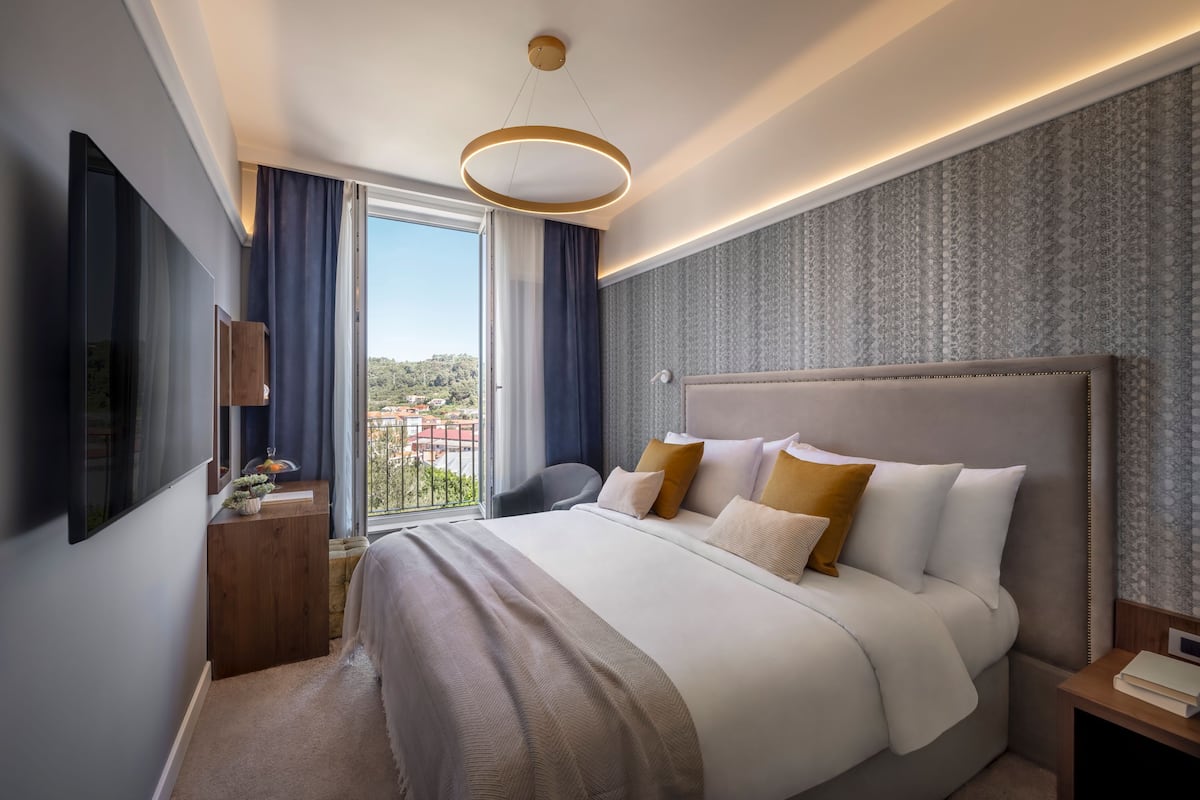 A modern hotel room with a large bed, a TV, a desk, and a view of the city.