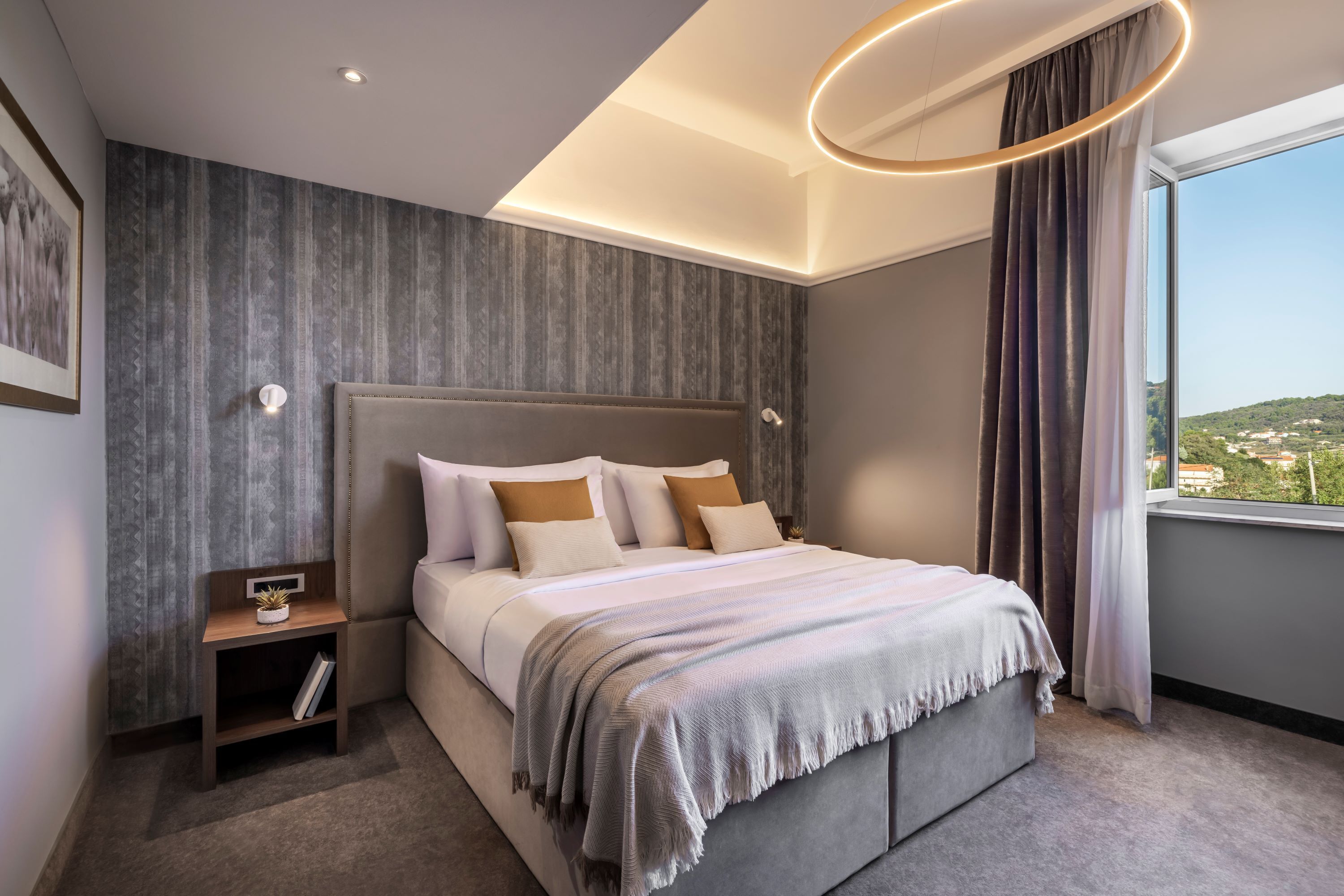 A modern bedroom with a large bed, a nightstand, a wall-mounted light, and a window with curtains.