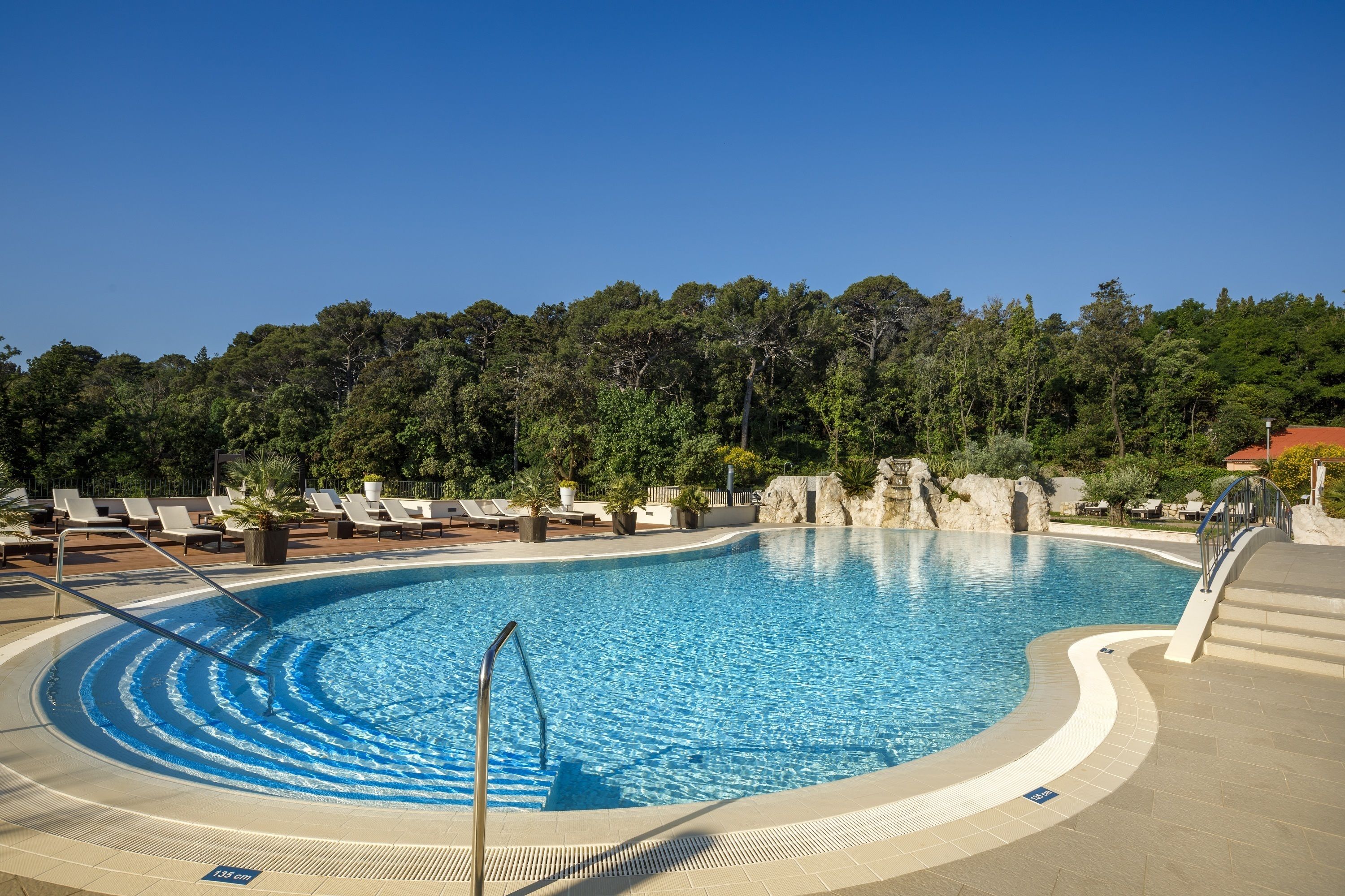 Piscina com borda curva, rodeada por cadeiras de descanso e um cenário arborizado.