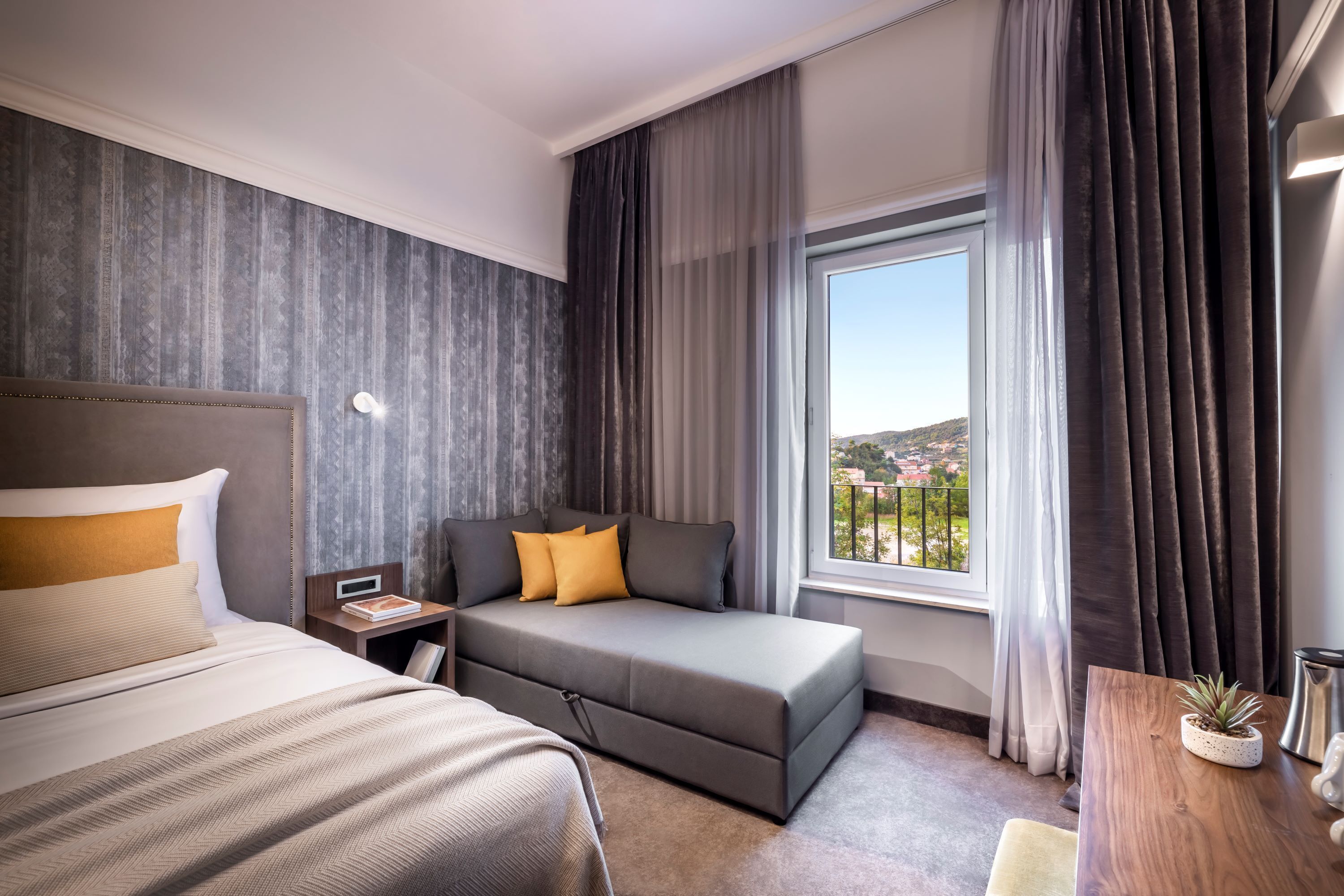 Een moderne hotelkamer met een bed, een sofa, een klein tafeltje met een plant en een raam met gordijnen.