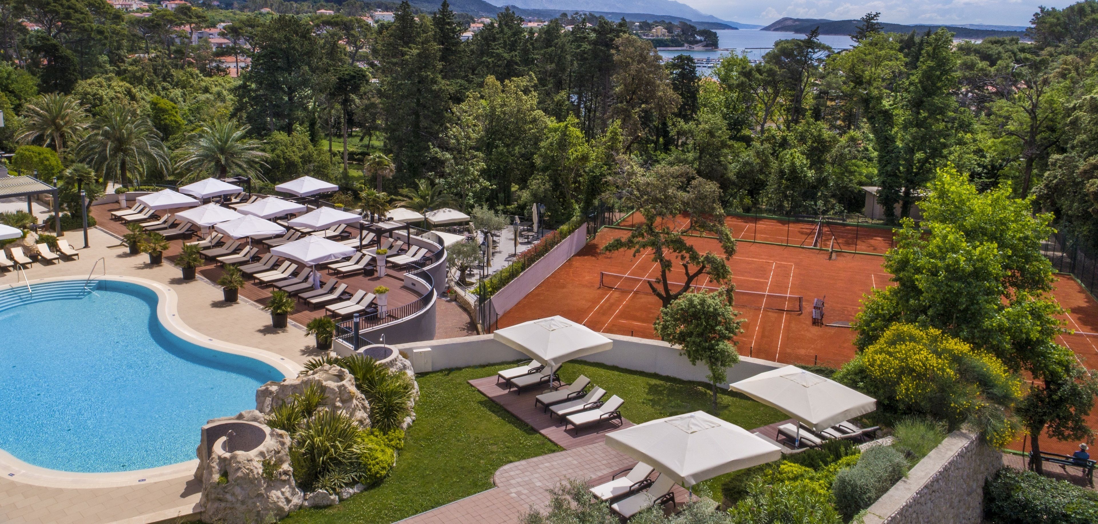 Un complexe hôtelier doté d’une piscine, de chaises longues et d’un court de tennis, le tout entouré d’une végétation luxuriante.