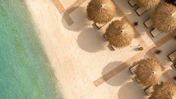 Luchtbeeld van een strandresort met helder turquoise water, een zandstrand en rijen rieten parasols en ligstoelen.