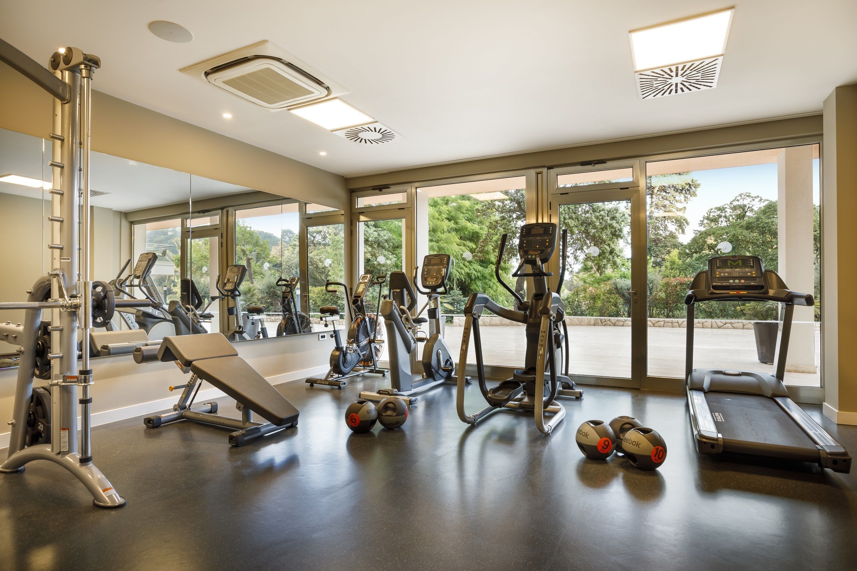 Un gimnasio moderno con diversos equipos de ejercicio, amplias ventanas y vistas al exterior con vegetación.