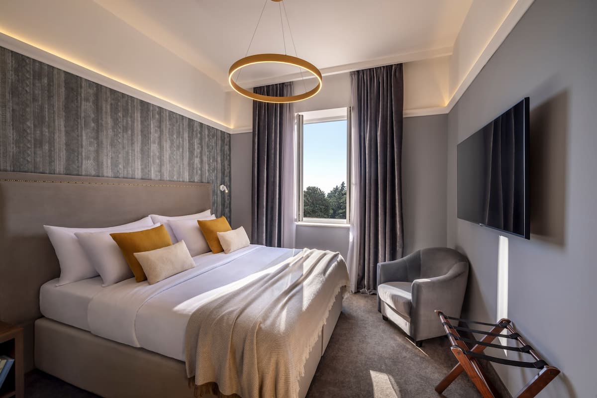 A modern hotel room with a large bed, a chair, a TV, and a window with curtains.