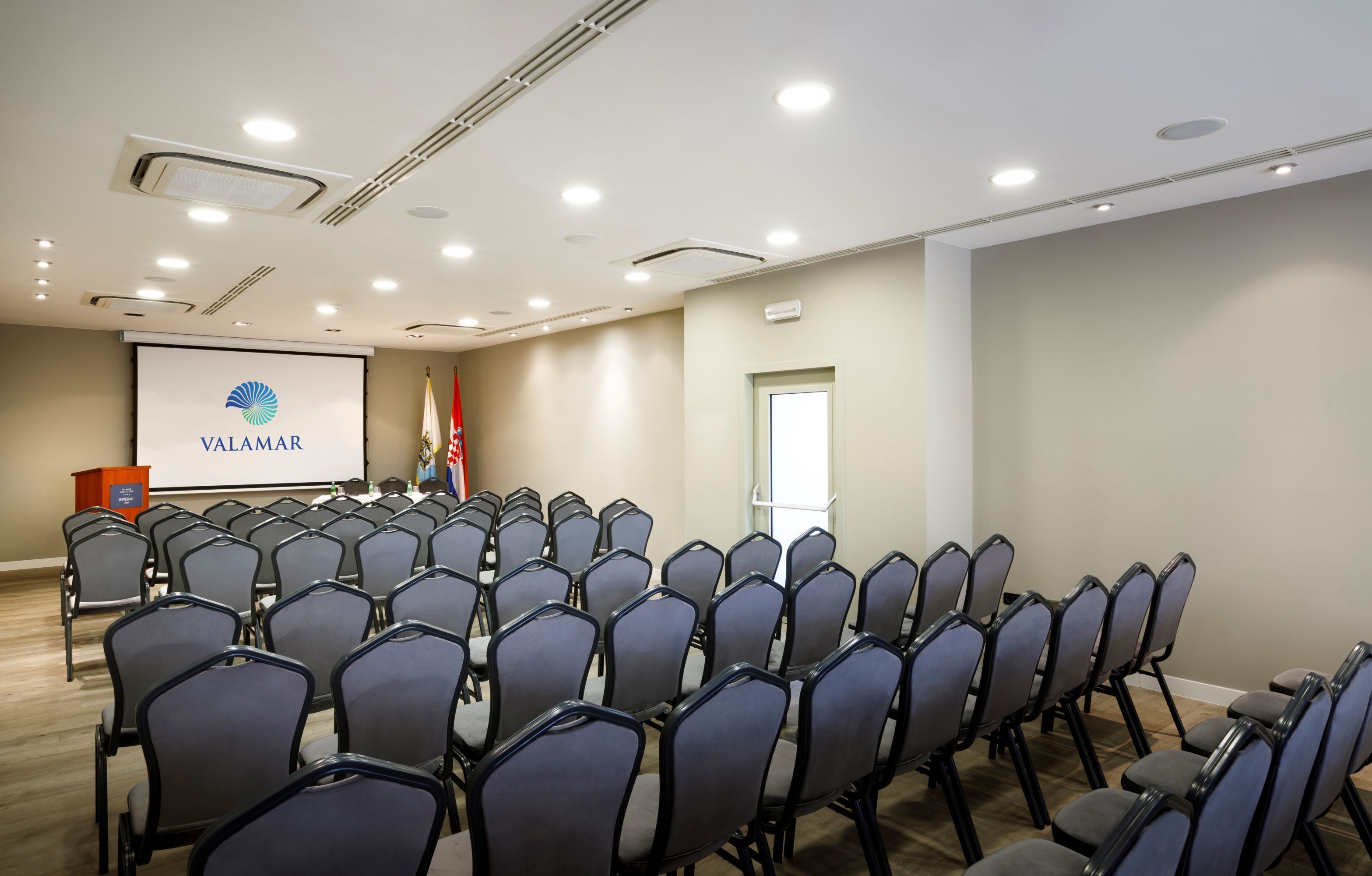 A conference room with rows of chairs, a podium, a projector screen displaying 'VALAMAR', and a flag.