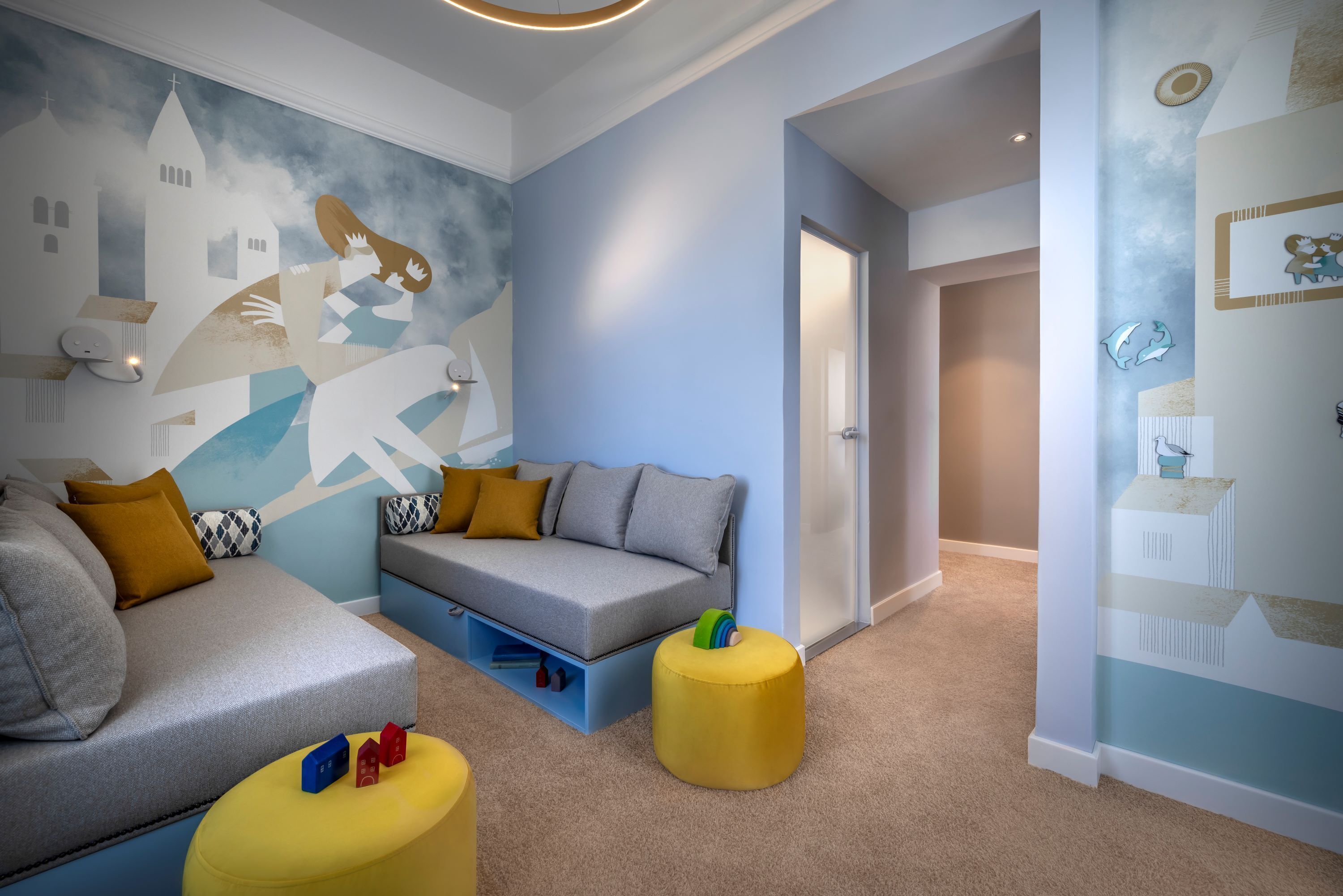 Un salon moderne avec un canapé gris, un canapé bleu et un pouf jaune. Un mur est décoré d’une fresque représentant une personne et des bâtiments.