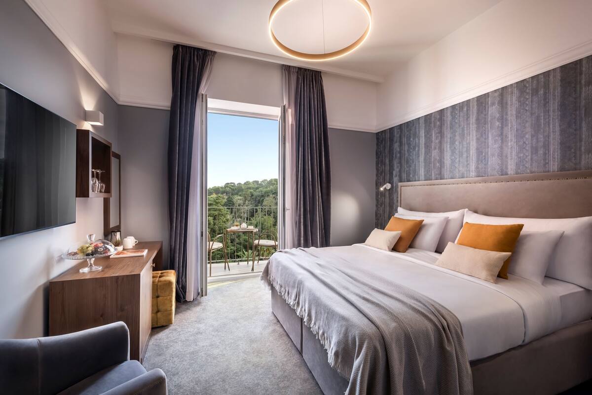 A modern hotel room with a large bed, a desk with a vase of flowers, a balcony with a view of greenery, and a large mirror.