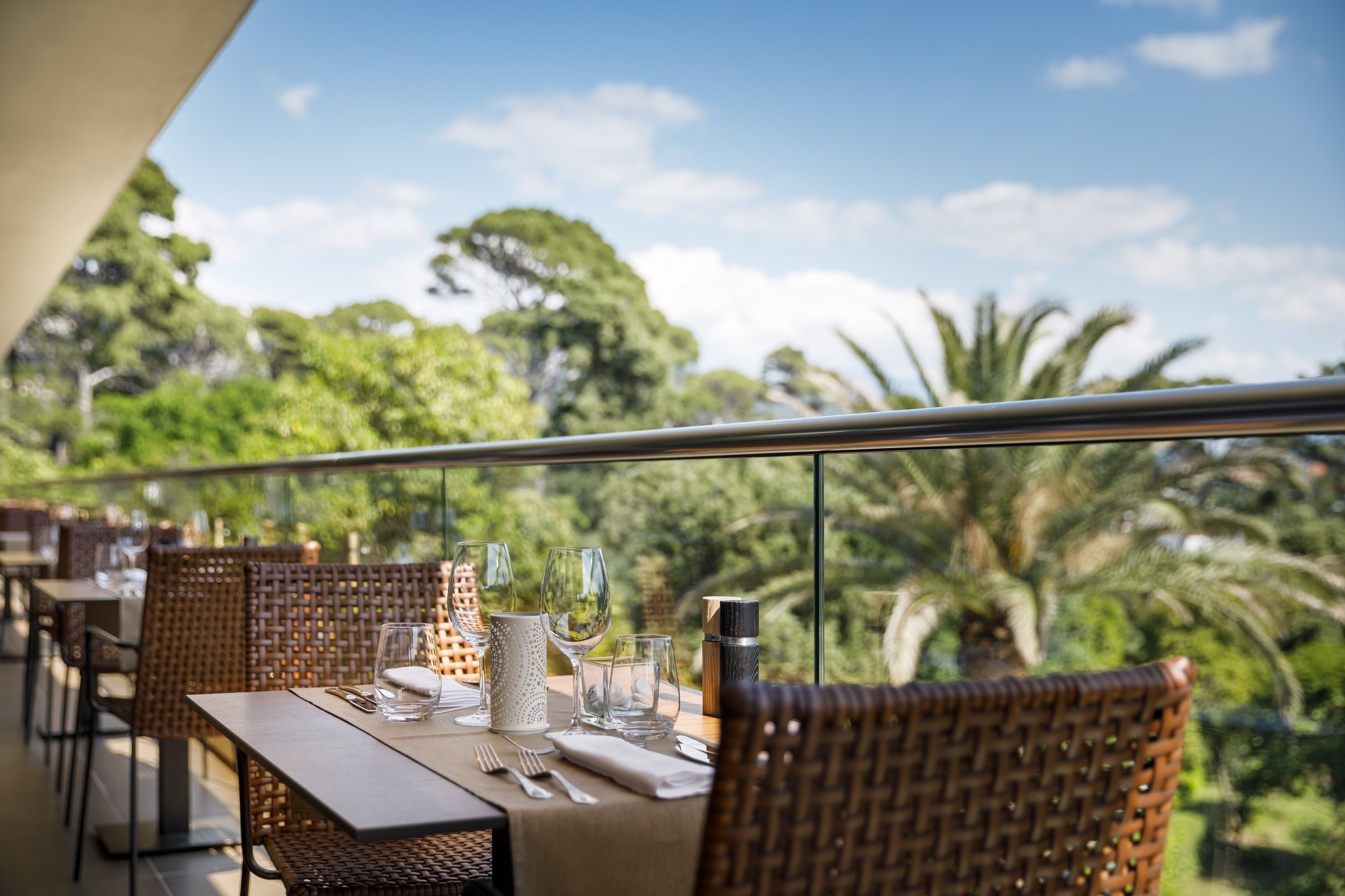 Un comedor con mesas dispuestas para dos, sillas de mimbre y vistas a abundante follaje.