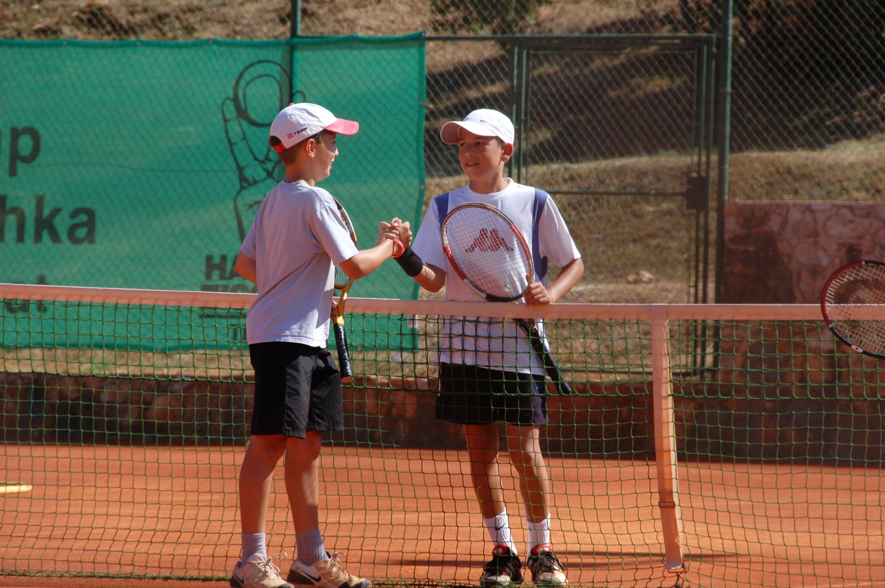 Zwei Personen geben sich auf einem Tennisplatz die Hand. Im Hintergrund ist ein grüner Zaun zu sehen, und einer der Spieler hält einen Tennisschläger.