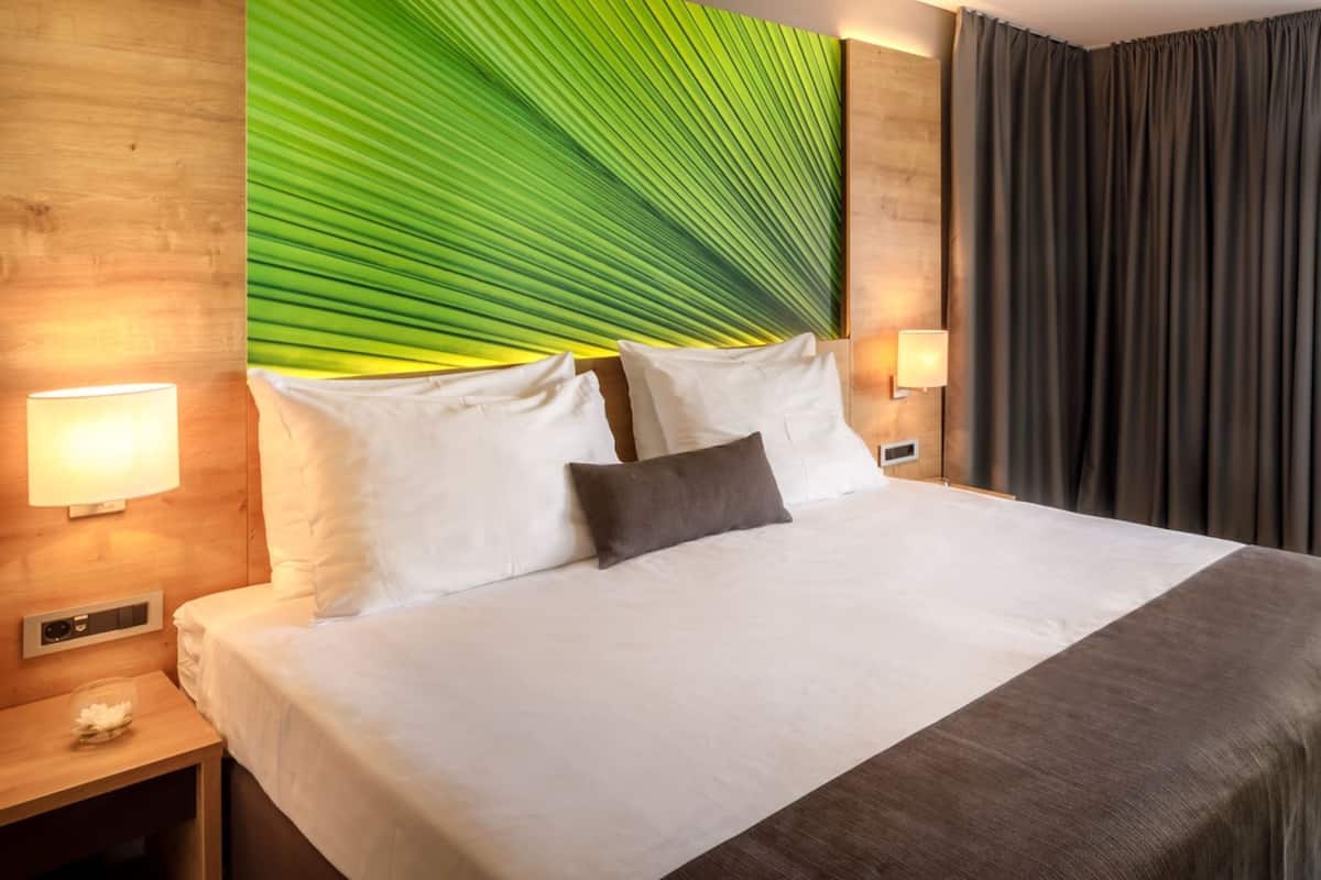 Une chambre d’hôtel avec un grand lit, deux lampes de chevet, une tête de lit en bois ornée d’un motif de feuilles vertes et un rideau foncé.
