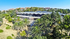Een modern hotel met een terras op het dak, omringd door palmbomen en groen.