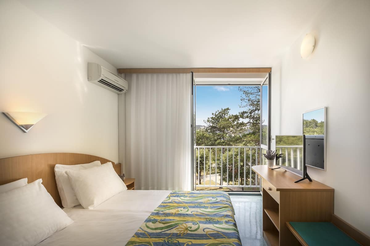 Une chambre d’hôtel avec un lit, un bureau avec un ordinateur, une télévision et un balcon donnant sur la verdure.