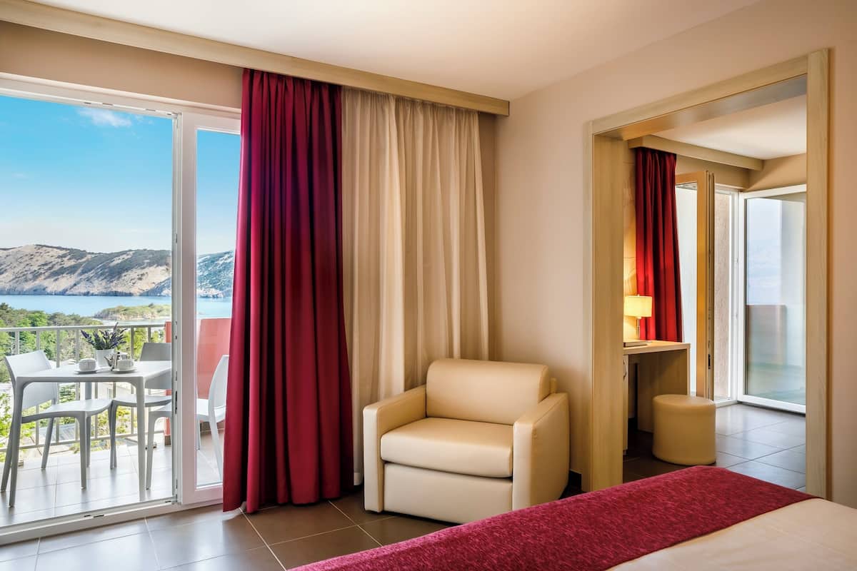 Ein Hotelzimmer mit Balkon, einem Sessel, einem Bett und einem Schreibtisch mit Lampe.