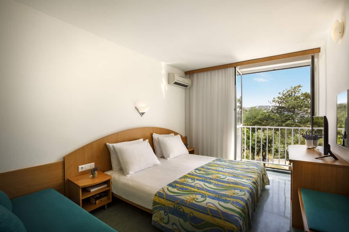 Une chambre d’hôtel avec un grand lit, un bureau, une chaise, une table de chevet et une fenêtre donnant sur un paysage verdoyant.