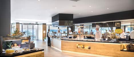 A modern kitchen area with chefs preparing food, a buffet counter with various dishes, and a view of the city through large windows.