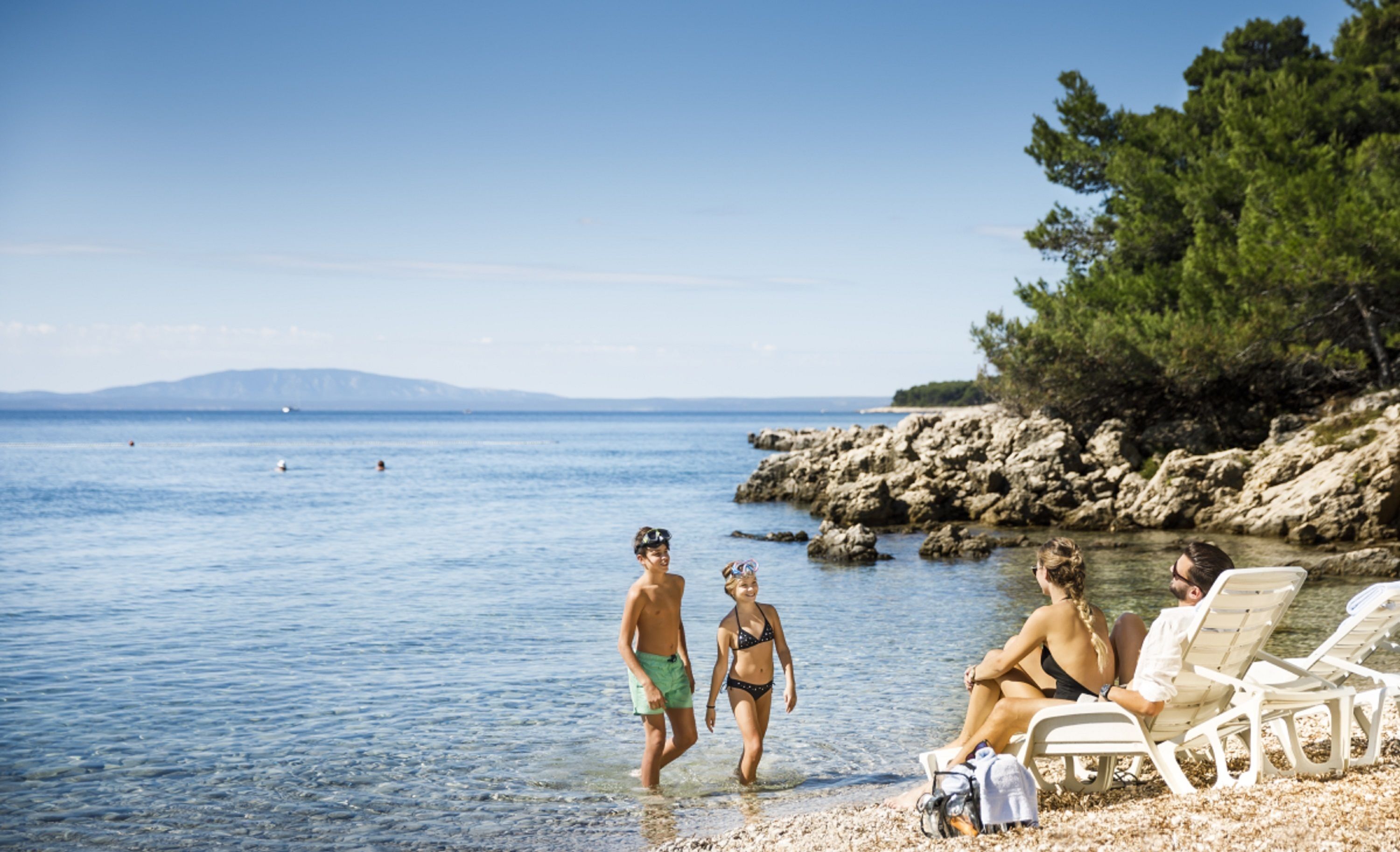 A beach scene with clear water, rocky shore, and a few people.