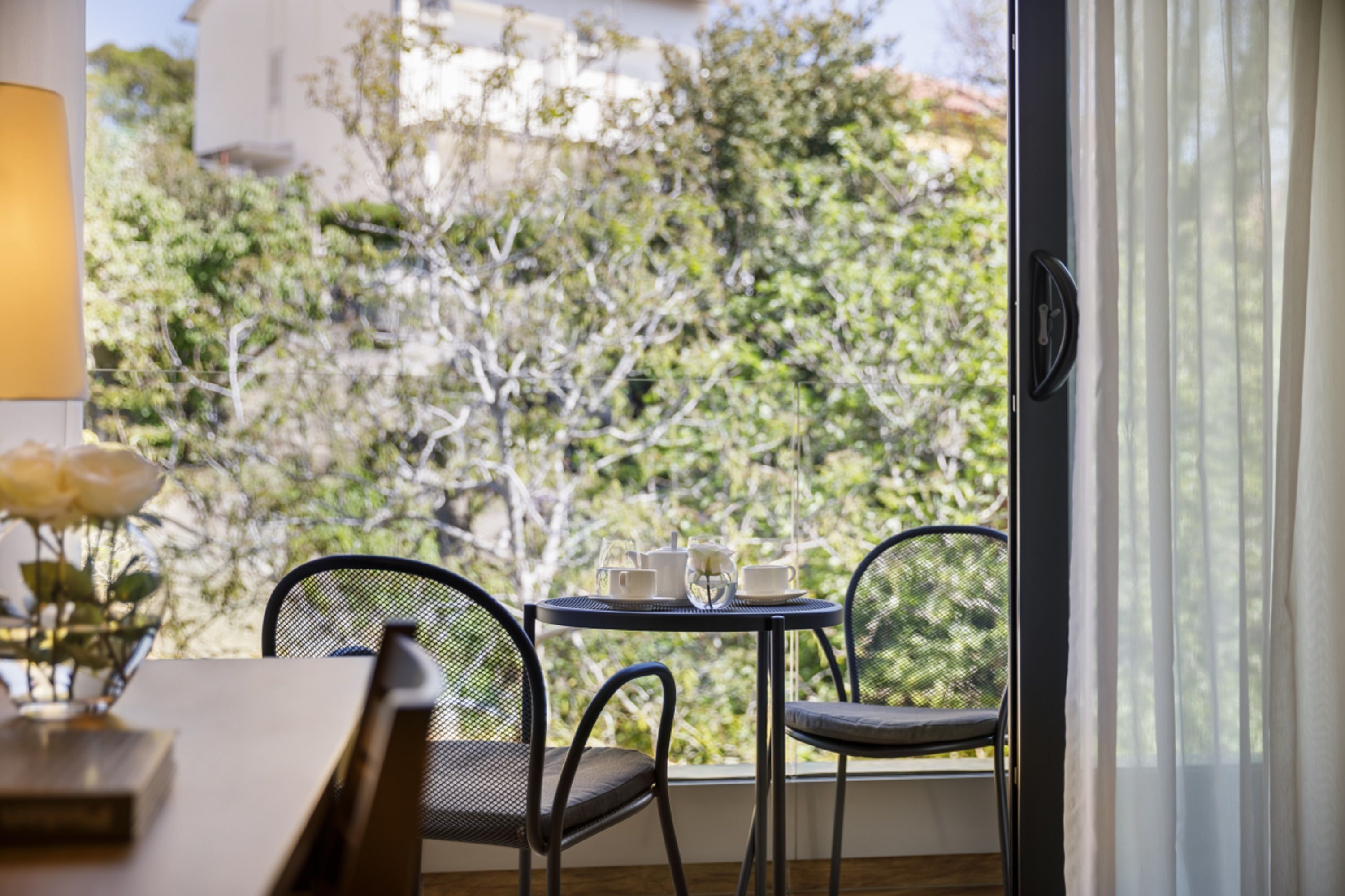 A room with a table and chairs near a window, a vase with flowers, and a view of trees and buildings outside.