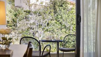 A room with a table and chairs near a window, a vase with flowers, and a view of trees and buildings outside.