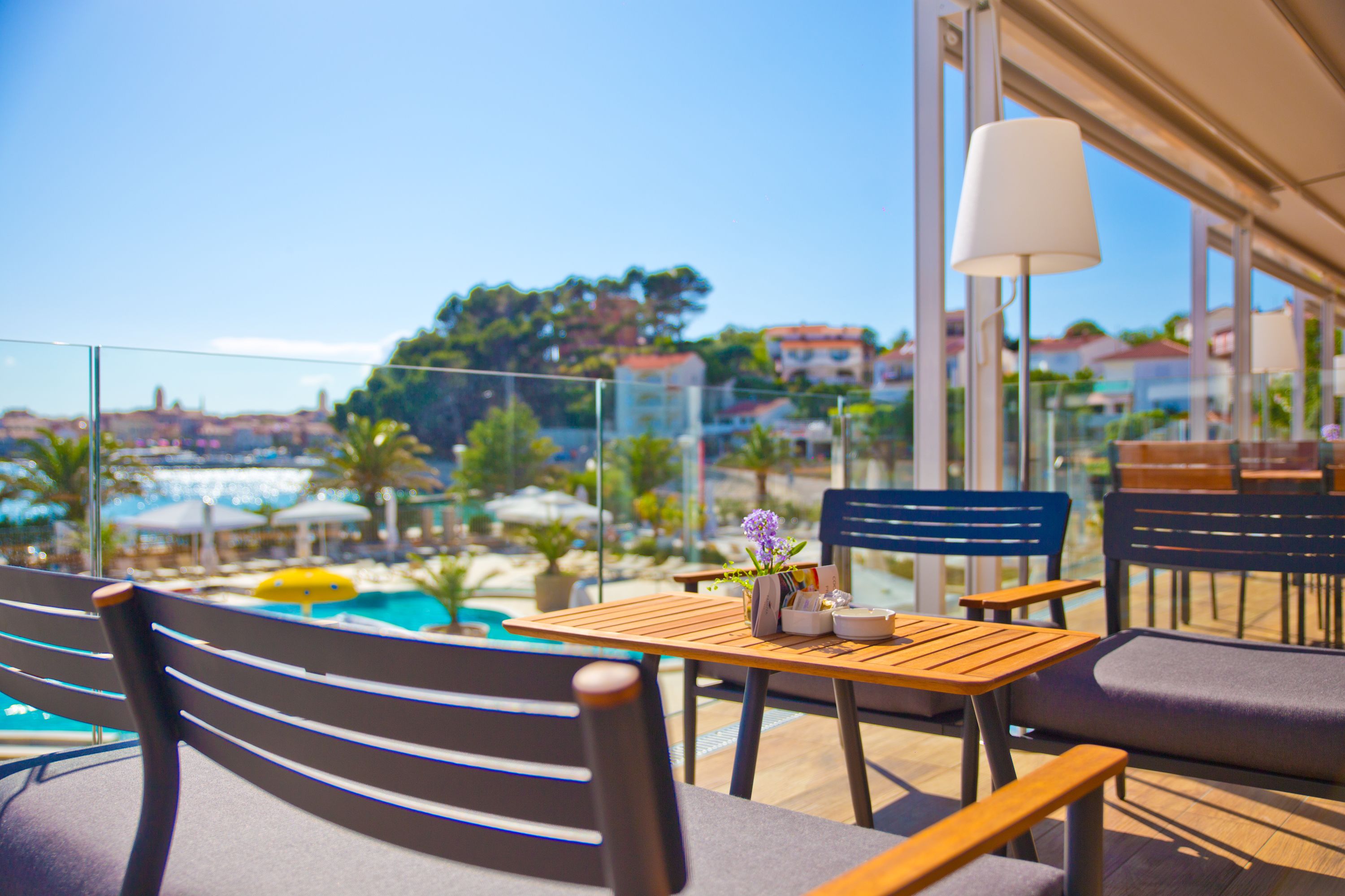 A terrace with a wooden table and chairs, a lamp, and a view of a pool and buildings in the background.