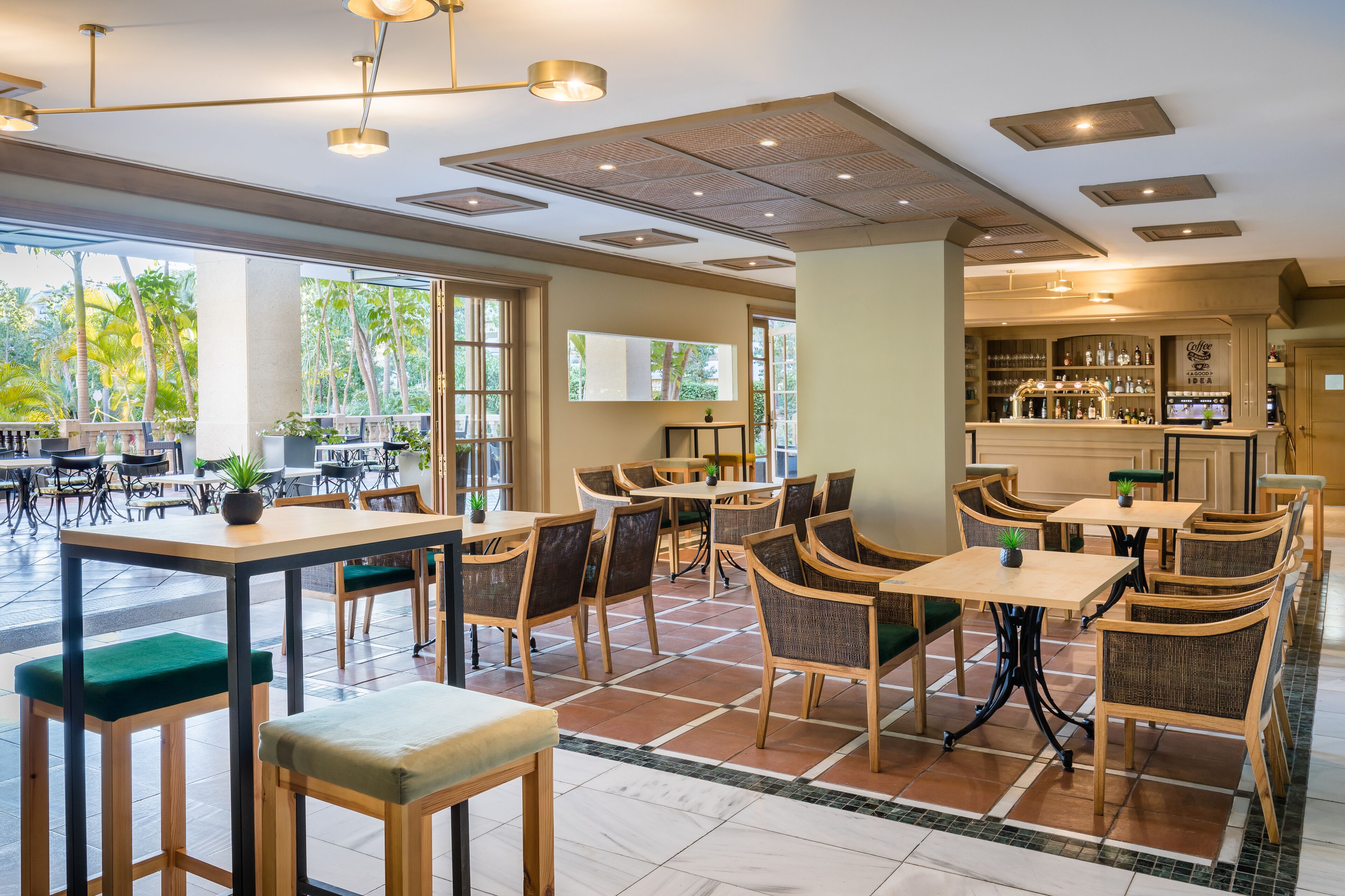 Een ruime, overdekte eetruimte met meerdere tafels en stoelen, grote ramen met uitzicht op palmbomen, en een bar met flessen op de planken.
