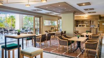 Een ruime, overdekte eetruimte met meerdere tafels en stoelen, grote ramen met uitzicht op palmbomen, en een bar met flessen op de planken.