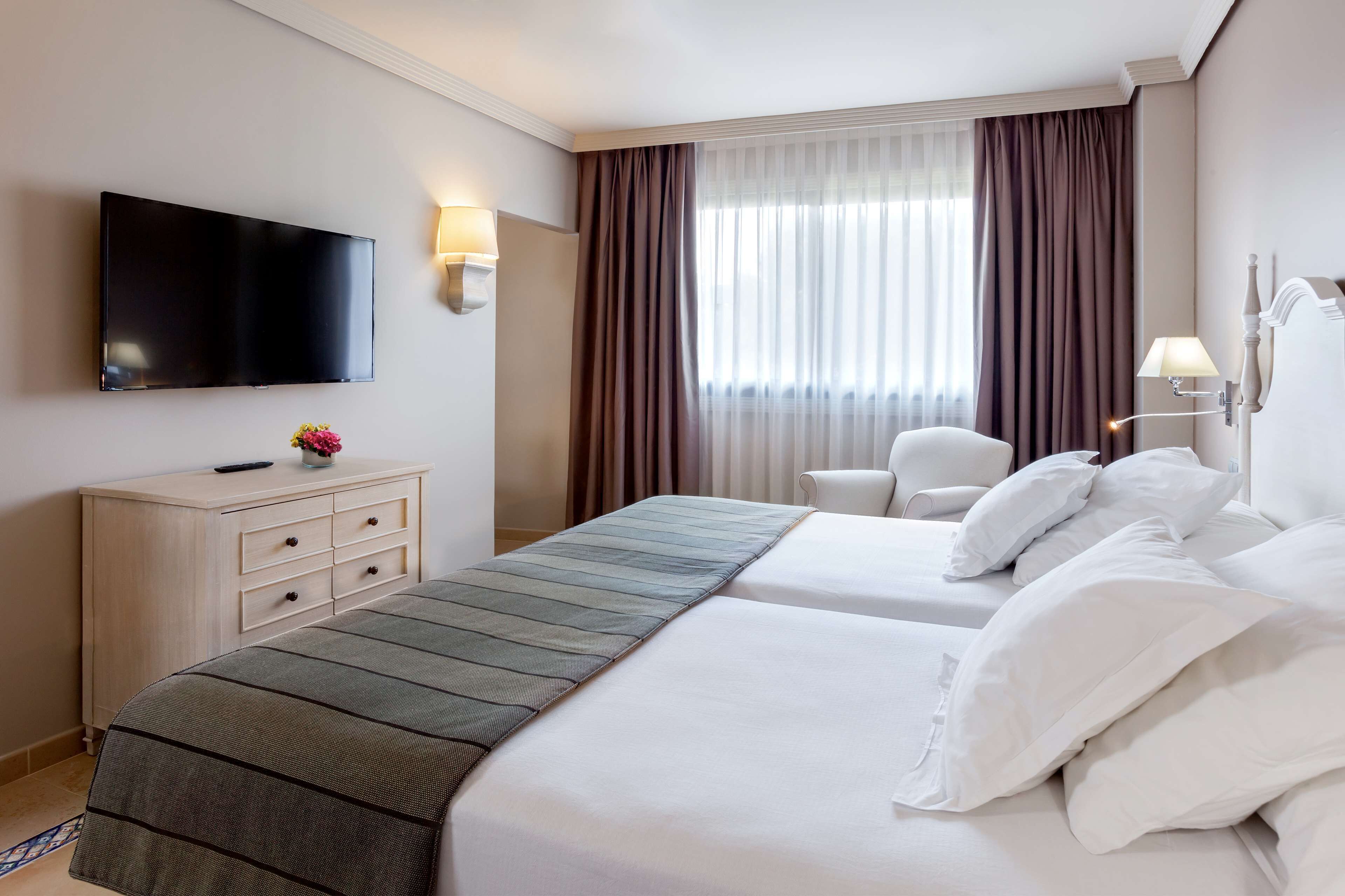 A hotel room with a flat-screen TV, a desk with drawers, a bed with white linens, and a chair.