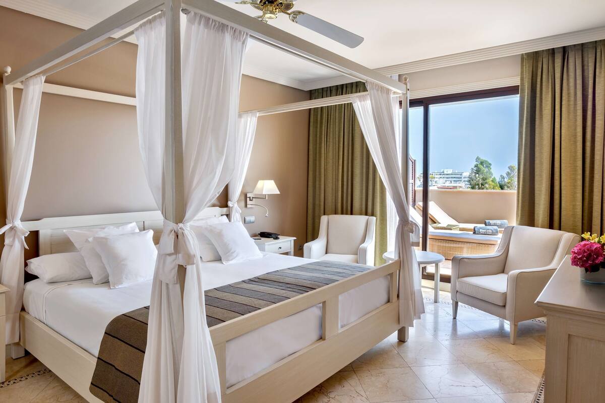 Una cama con dosel, ropa de cama blanca, una mesita de noche con lámpara, una silla y vistas a la ciudad a través de amplios ventanales.