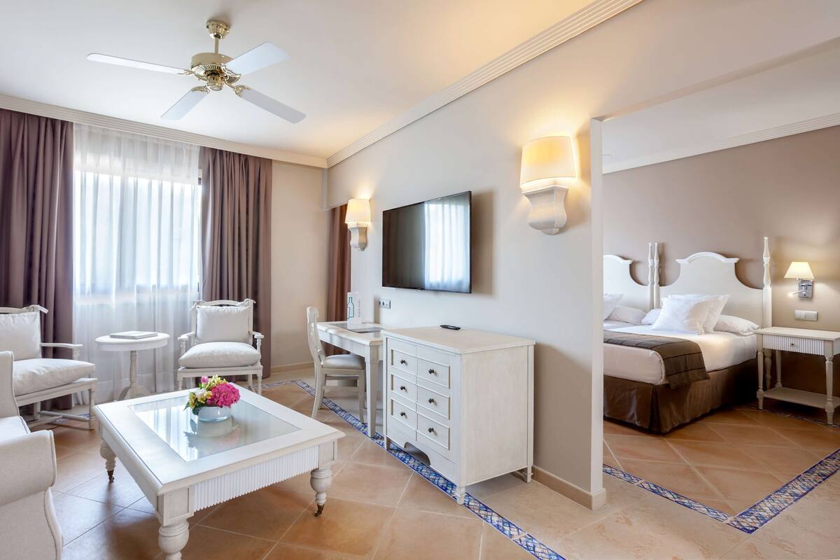 Una habitación de hotel con una cama, un escritorio, una silla, una mesa de centro, un televisor y un ventilador de techo.