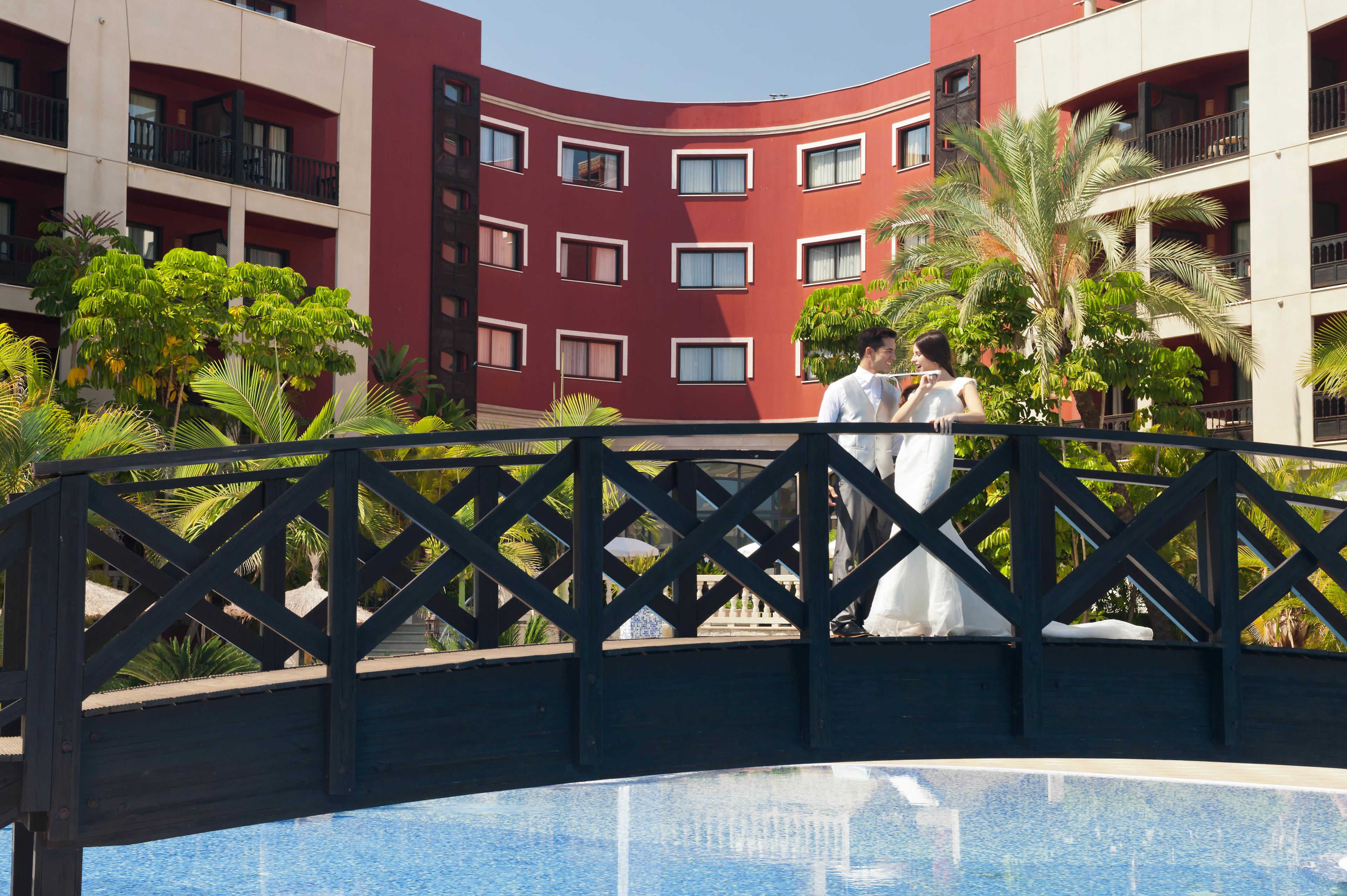 Un hotel con fachada roja curva, balcones y un área de piscina con palmeras.