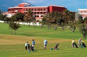 A golf course with a red building in the background, palm trees, and people playing golf.