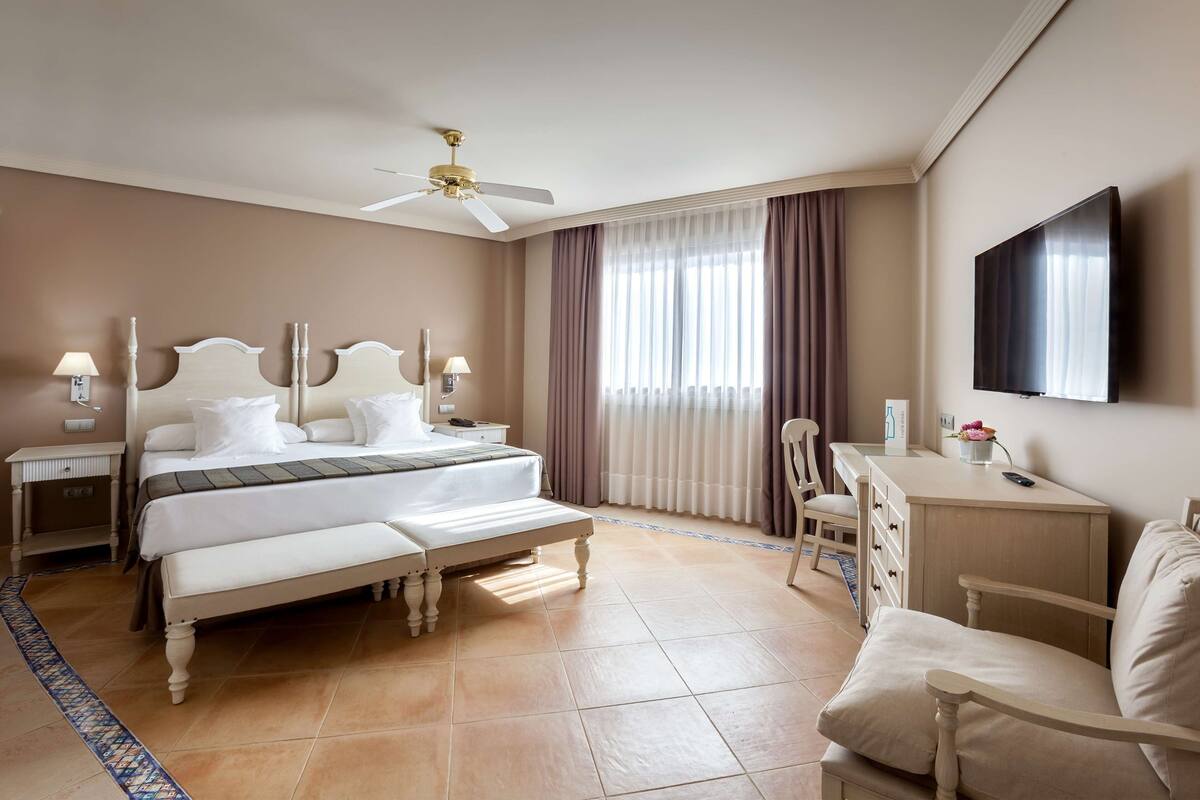 Habitación de hotel con cama, mesitas de noche, escritorio, silla, televisor y ventilador de techo.