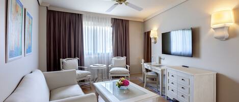A hotel room with a white sofa, a coffee table with a vase, a TV, a white cabinet, and a ceiling fan.