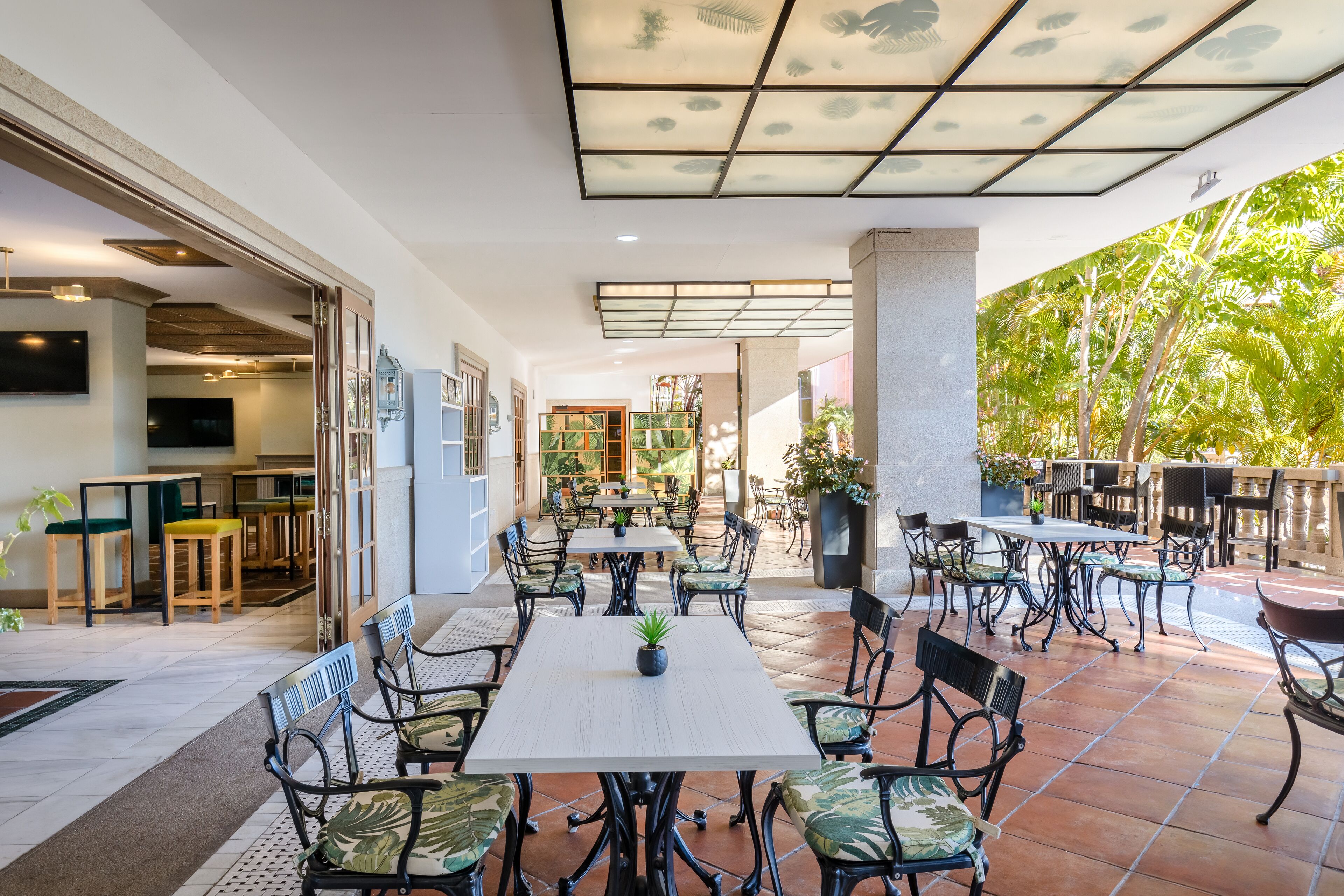 Un patio al aire libre con varias mesas y sillas, un techo con diseño de hojas y vistas a un jardín con árboles.