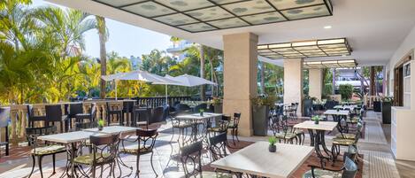 An outdoor patio area with tables and chairs, surrounded by palm trees and plants.