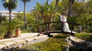 A couple on a bridge over a pond in a lush garden with palm trees and a building in the background. - Barceló Marbella (Marbella)