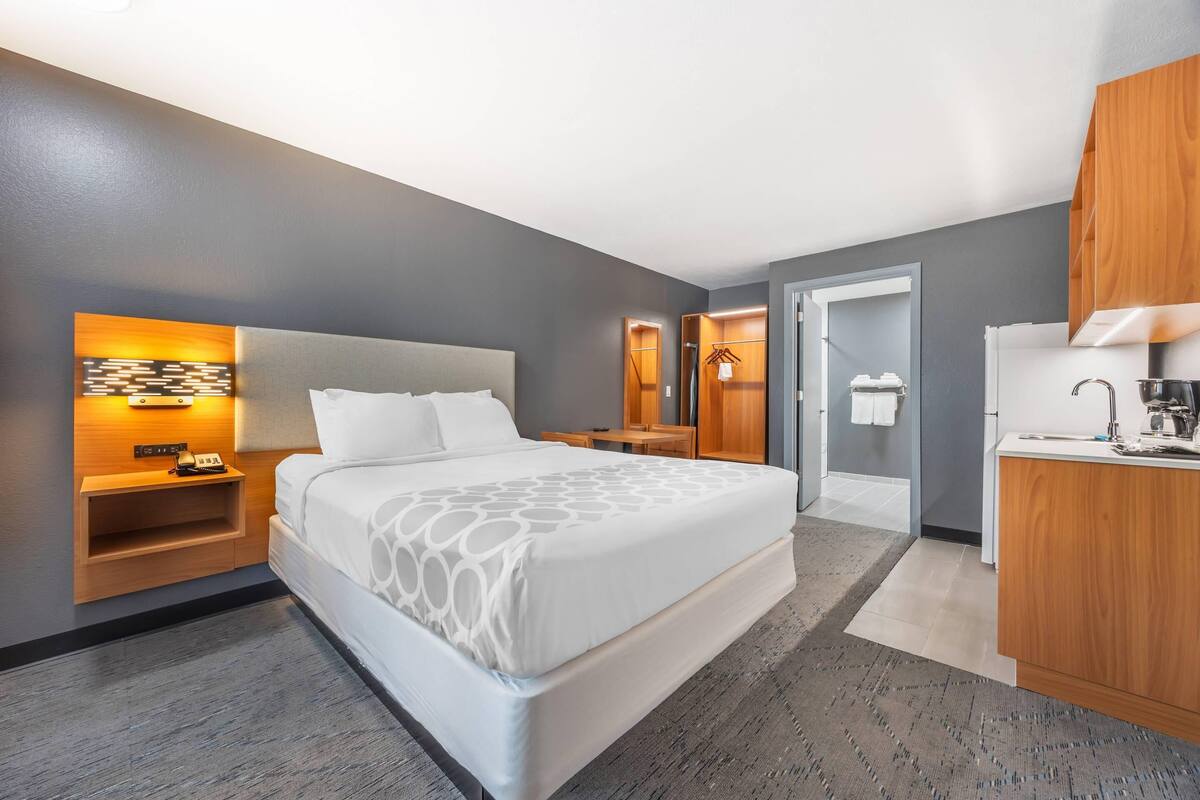A modern hotel room with a large bed, bedside tables, a built-in desk, and a small kitchenette.