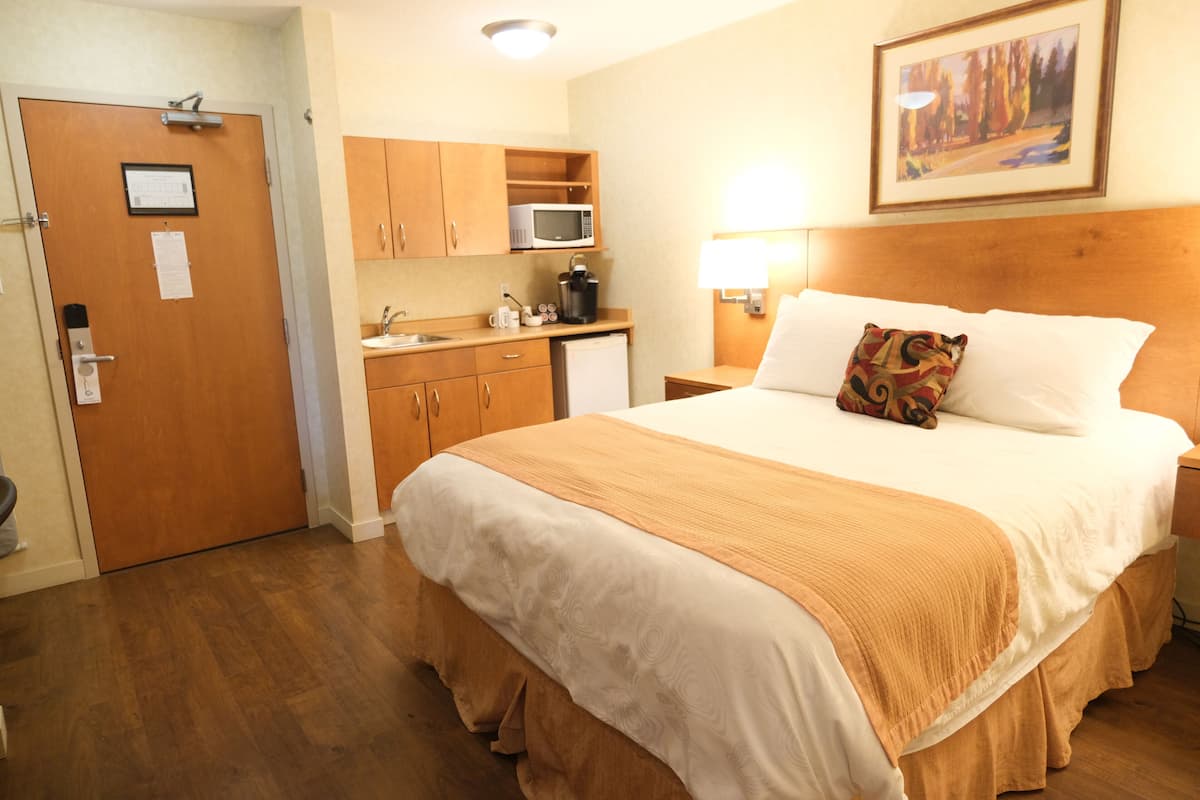 Habitación de hotel con cama, mesita de noche, lámpara, microondas, cafetera y pequeña kitchenette.