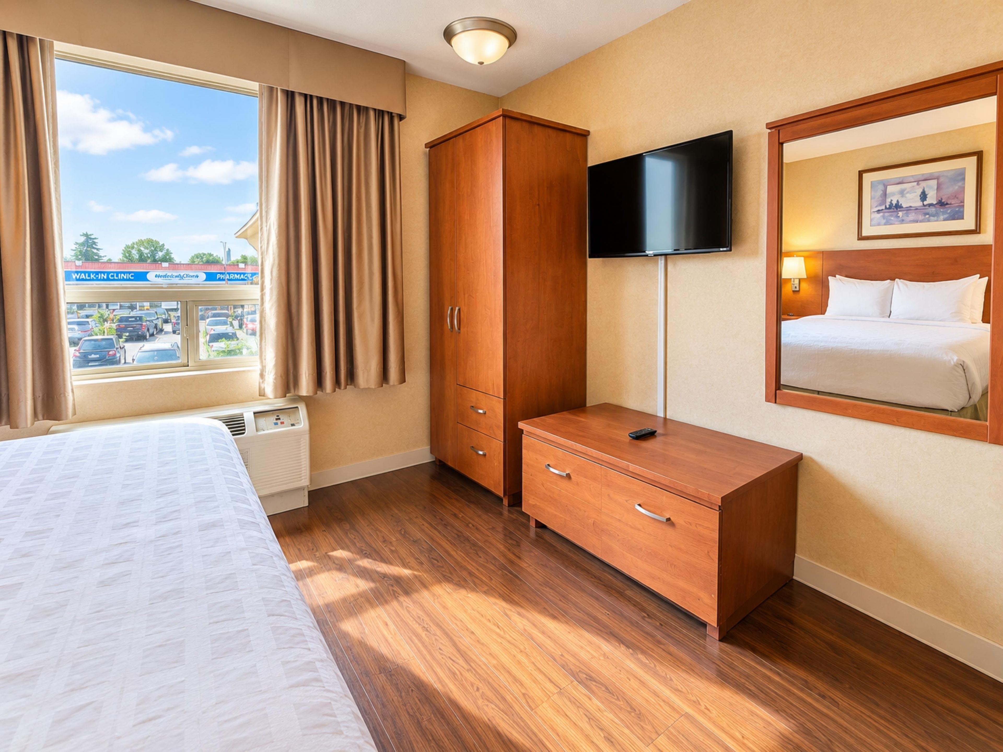 A hotel room with a bed, a wooden wardrobe, a television mounted on the wall, and a window with curtains.